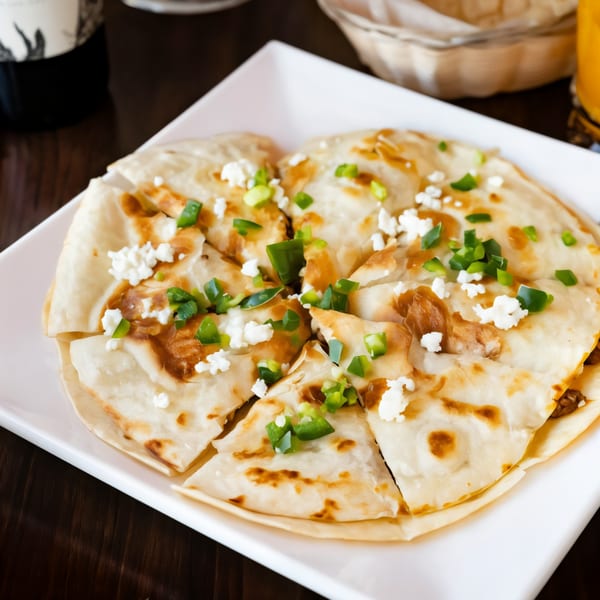A round, cut quesadilla topped with green onions and white cheese on a white plate.